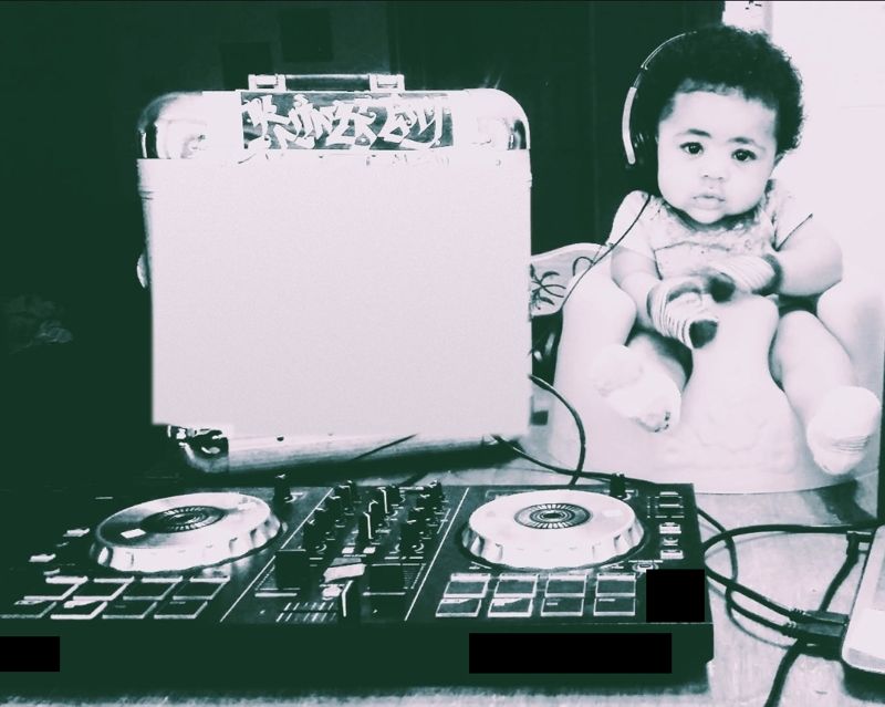 A baby wearing headphones at a DJ controller, beside a case that reads That’s My DJ
